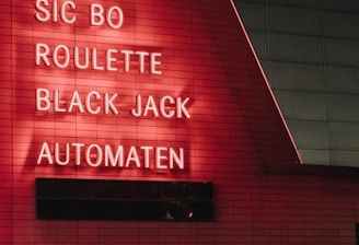 Red neon letters are mounted on a dark tiled wall, displaying the words Poker, Sic Bo, Roulette, Black Jack, and Automaten.