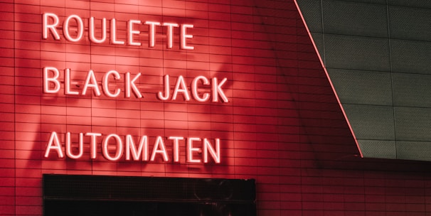 Red neon letters are mounted on a dark tiled wall, displaying the words Poker, Sic Bo, Roulette, Black Jack, and Automaten.