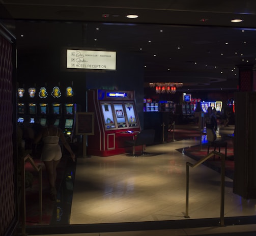 A dimly lit casino with several rows of slot machines, some displaying bright graphics and lights. There are a few people scattered throughout the area, engaging with the machines. The floor is reflective and polished, adding to the ambiance, while the ceiling is dotted with numerous small lights.
