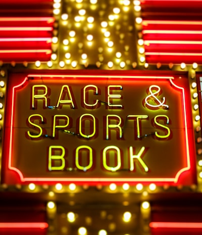 Bright neon lights on a marquee sign display the words 'RACE & SPORTS BOOK'. The sign is surrounded by rows of yellow and white lights against a red background, evoking a vibrant, energetic atmosphere typically associated with casinos or entertainment venues.